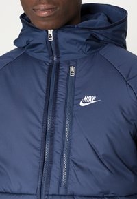 Jūrininkų stiliaus pūkinė striukė su aukštu apykakle ir reguliuojama kapišonu. Yra dvigubi priekiniai užtrauktukai ir balta "Nike" logotipas kairėje krūtinės pusėje.