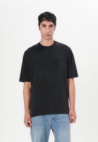 Schwarzes Baumwoll-T-Shirt mit Rundhalsausschnitt und kurzen Ärmeln. Hat ein dezentes Ton-in-Ton-Logo auf der linken Brust. Leicht entspannter Schnitt.