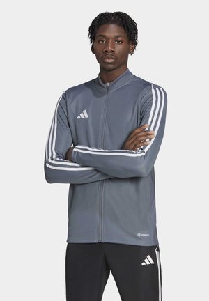 Graue Sportjacke mit durchgehendem Reißverschluss, weißen Drei-Streifen-Akzenten an den Ärmeln und Logo. Glatte Textur, taillierte Passform und kontrastierende Elemente.