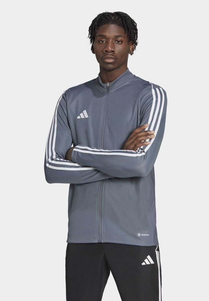 Graue Sportjacke mit durchgehendem Reißverschluss, weißen Drei-Streifen-Akzenten an den Ärmeln und Logo. Glatte Textur, taillierte Passform und kontrastierende Elemente.
