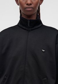 Personne aux cheveux tressés portant une veste noire zippée avec un petit logo Carhartt WIP en forme de cœur sur la poitrine.