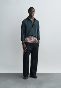 Jeune homme debout contre un mur gris clair, portant une chemise vert foncé, une chemise à carreaux rouge et blanc superposée, un pantalon noir et des chaussures noires.