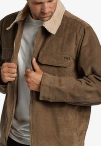 Chaqueta de pana marrón con un collar de piel sintética beige, cremallera frontal y dos bolsillos en el pecho. Tejido texturizado con líneas verticales.
