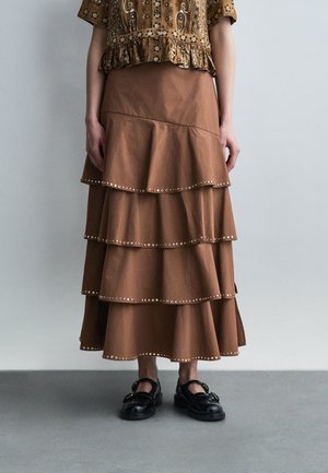 Brauner, gestufter Rock mit Metallnieten entlang der Kanten, getragen mit schwarzen Schnallenschuhen und gemustertem Kurzarmbluse, stehend auf grauem Boden.