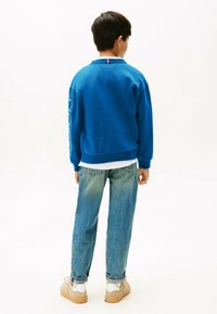 Sudadera azul con puños acanalados, con un forro blanco en el cuello y bordado en los laterales. Lleva puesta con jeans azul claro y zapatillas beige.