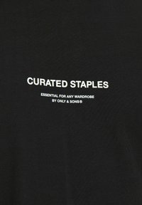 Svart bomull t-shirt med vit text som säger "KURATERADE STAPELVAROR, AVGÖRANDE FÖR VARJE GARDEROB AV ONLY & SONS," med en mjuk textur.