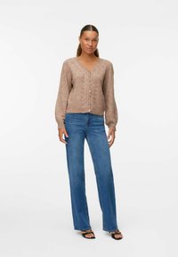Cardigan beige à texture avec un col en V, des boutons sur le devant et des manches longues, associé à un jean bleu à jambes larges et des sandales noires.
