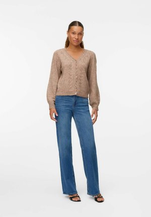 Cardigan beige à texture avec un col en V, des boutons sur le devant et des manches longues, associé à un jean bleu à jambes larges et des sandales noires.