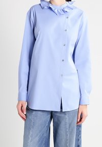 Camicia azzurro chiaro con colletto arricciato, maniche lunghe e taglio dritto. Presenta bottoni metallici lungo la parte anteriore e un orlo spaccato.