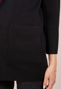 Svart stickad cardigan med en framficka, långa ärmar och en texturerad tygfinish. Synlig sömnad och kantdetaljer finns.