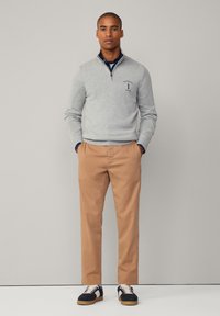 Sudadera gris con cremallera y acentos en azul marino, chinos beige y zapatillas oscuras con cordones crema. Presenta un logo en la sudadera. Diseño sencillo.