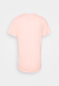 Camisa rosa claro de manga corta mostrada desde la parte posterior con un dobladillo redondeado sobre un fondo blanco liso.