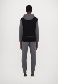 Man som står med ryggen mot kameran, iförd svart och grå huvtröja samt grå joggers med vita sneakers, framför en enfärgad bakgrund.