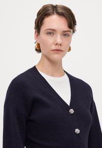 Σκουρό μπλε κασκόλ φτιαγμένο από μαλακό ύφασμα, με V λαιμόκοψη, μακριά μανίκια και δύο μεταλλικά λουλουδάτα κουμπιά μπροστά.