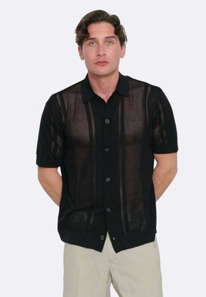 Uomo dalla pelle chiara e occhi verdi che indossa una camicia nera semi-trasparente a maniche corte abbottonata e pantaloni beige su uno sfondo semplice.