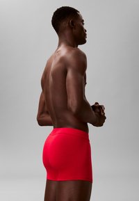 Boxer ajustados de color rojo hechos de tela suave. Presentan una cintura lisa y un corte a mitad de muslo. El modelo está mostrado de perfil.