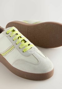White leather and suede sneakers with yellow laces, textured brown rubber sole, and a side stripe in a matching yellow accent.