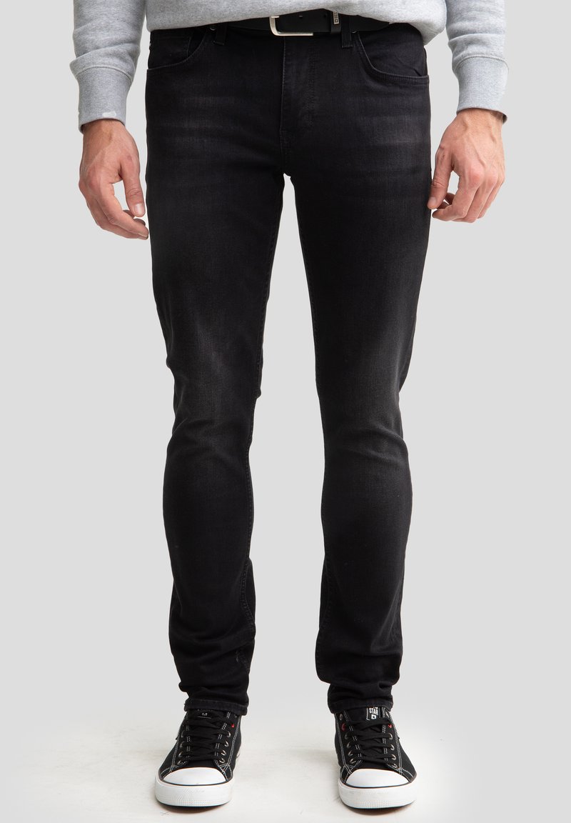 Mustad slim-fit teksased, mis on valmistatud denimist, omavad sujuvat tekstuuri ja peent kulumist. Need on sobitatud mustade kõrgemate spordijalanõudega, millel on valged tallad.