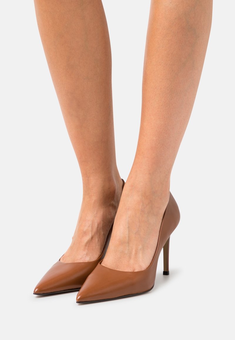 Chaussures à talons hauts en cuir marron avec un bout pointu, une texture lisse et une silhouette élégante, montées sur des talons aiguilles fins.