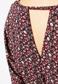 Blus med blommönster som har en djup V-ringning, svart bakgrund med röda och vita blommor, och ett urklipp på baksidan.