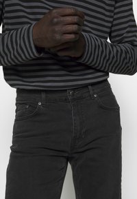 Jeans de mezclilla negra con un diseño de cinco bolsillos, que presentan un cierre de botón y cremallera; combinados con una blusa de manga larga de rayas grises.