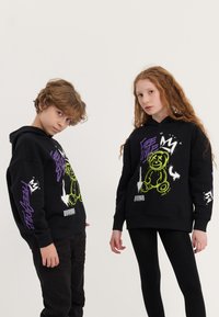 Os hoodies pretos apresentam designs gráficos coloridos, incluindo um urso e texto nas cores roxa e verde. Usados com leggings e calças pretas.
