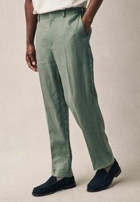 Hellgrüne Leinenhosen mit geradem Schnitt, Seitentaschen und einem knöpfbaren Bund, kombiniert mit marineblauen Slippern und grauen Socken.