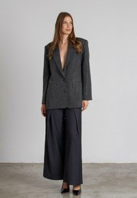 Grijze gestructureerde blazer met een diep V-hals, twee knopen en textielstof, gecombineerd met donkere, wijde broekspijpen en zwarte hakken.