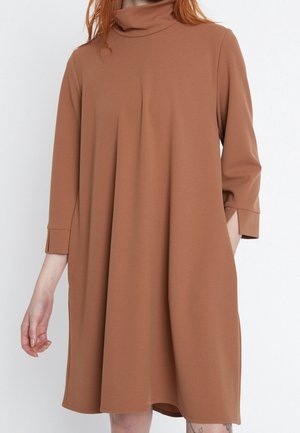 Femme portant une robe marron ample, longueur genoux, avec des manches 3/4 et un col roulé haut et décontracté, sur fond blanc uni.
