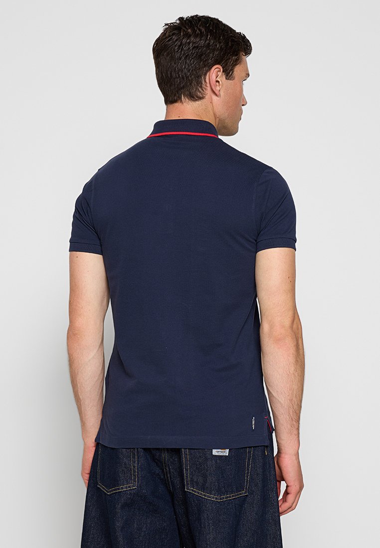 Dunkelblaues Poloshirt aus Baumwolle, mit kurzen Ärmeln und einem kontrastierenden roten Kragen, logo am unteren Saum und seitlichen Lüftungsöffnungen.