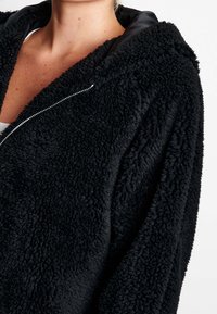 Czarna, puszysta bluza z kapturem wykonana z miękkiego, teksturowanego materiału, z zamkiem z przodu i kapturem wyłożonym satyną. Bez widocznych wzorów lub zdobień.