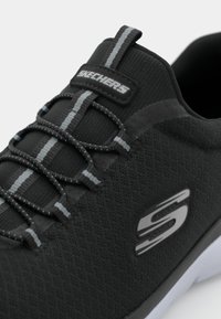 Scarpa da ginnastica nera con parte superiore testurizzata, lacci grigi a contrasto e logo Skechers sul lato. Design flessibile con suola ammortizzata.