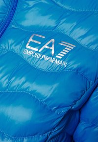 Jasnoniebieska pikowana kurtka z logo „EA7 Emporio Armani” w srebrnych literach na piersi, obok zamka błyskawicznego.