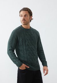 Mørk grønn cable-knit genser har teksturerte mønstre, rund hals, og ribbede mansjetter, sammen med mørke bukser.