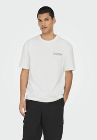 T-shirt en coton blanc à manches courtes, avec un petit logo en texte noir sur la poitrine gauche. Coupe décontractée, texture douce.