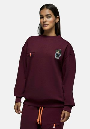 Femme portant un sweat-shirt bordeaux et un pantalon assorti avec des cordons orange et des patchs décoratifs sur la poitrine et la poche.