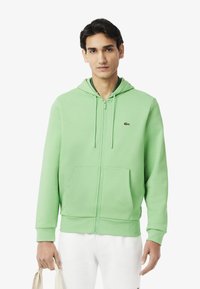 Haut de sweatshirt zippé vert clair avec une poche avant et une capuche à cordon. Matériau doux, poignets côtelés et un petit logo de marque sur la poitrine.