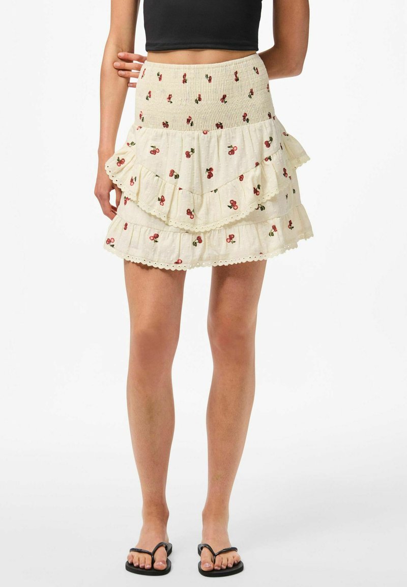 Jupe à volants superposés en crème avec des motifs de cerises, dotée d'une taille smockée et d'une bordure en dentelle au bas. Fabriquée en tissu léger.