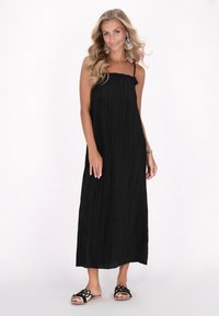 Schwarzes, plissiertes Maxikleid mit dünnen Trägern und einem Rüschenausschnitt. Das Modell trägt schwarze, verzierten Rutschsandalen. Strukturiertes Material.