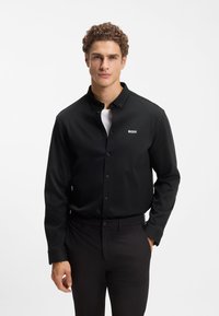 Μαύρο πουκάμισο με κουμπιά, με λεία υφή και γιακά, που φέρει μικρό λευκό λογότυπο «BOSS» στο στήθος, συνδυασμένο με σκουρόχρωμο παντελόνι.