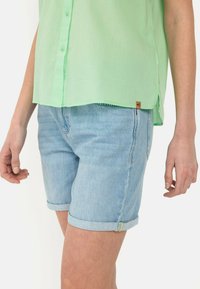Hellgrünes Kurzarmhemd mit Knopfleiste und einem Etikett am Saum, kombiniert mit hellblauen Jeansshorts mit umgeschlagenem Saum.