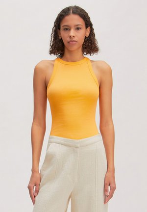 Young woman with curly hair wearing a sleeveless yellow top and high-waisted textured white pants, standing against a plain background.