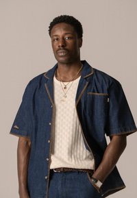 Denim short-sleeve shirt with contrast stitching, worn over a cream textured knit top. Accessories include layered necklaces and a watch.