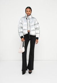 Chaleco de plumas plateado con cuello alto y cierre de cremallera, combinado con pantalones negros acampanados y un bolso pequeño rosa, junto con unos tacones negros de punta.