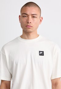 Cremefarvet bomulds-t-shirt med korte ærmer, med et sort Nike Air-logo patch på venstre bryst. Glat tekstur, regular fit.