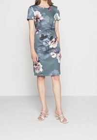 Robe à motif floral en teal, à manches courtes, longueur genoux. Tissu lisse avec des fleurs roses, bleues et blanches. Portée avec des talons ouverts de couleur claire.