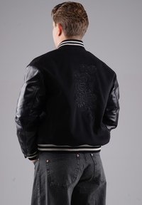 Schwarze Varsity-Jacke mit Lederärmeln, gesticktem Design auf dem Rücken, geripptem Kragen und Saum. Verfügt über horizontale weiße Streifen am Kragen.