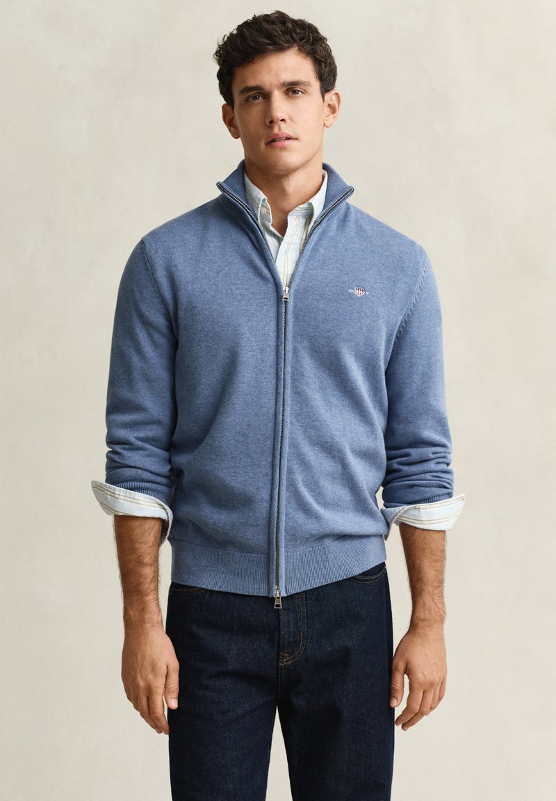 Uomo che indossa un maglione azzurro chiuso con cerniera sopra una camicia bianca con collo e maniche arrotolate, e jeans scuri, in piedi contro uno sfondo semplice.