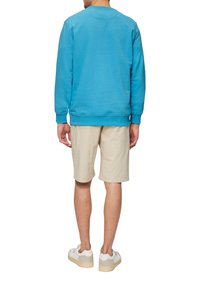 Blaues Sweatshirt mit langen Ärmeln und Rundhalsausschnitt, kombiniert mit beigen Shorts. Das Model trägt weiße Sneakers mit grauen Akzenten.