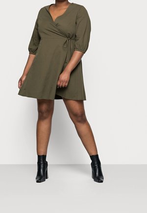 Personne portant une robe portefeuille vert olive à manches trois-quarts et des bottines noires à talons, sur un fond clair uni.